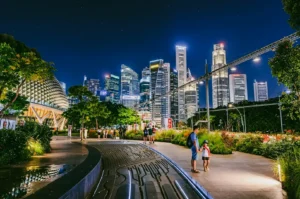 Eye-level wide-angle shot of Esplanade Park Singapore at night with illuminated Marina Bay skyline, landscaped pedestrian walkway, and visitors enjoying an urban green space, highlighting community interaction and vibrant public spaces.