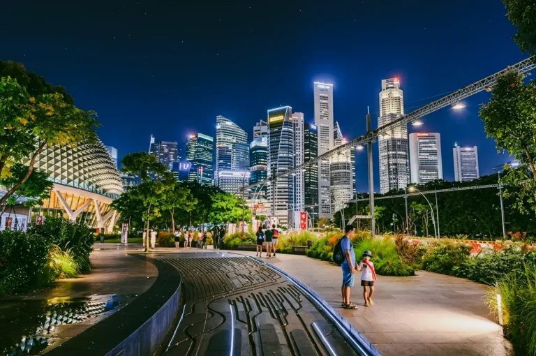 Eye-level wide-angle shot of Esplanade Park Singapore at night with illuminated Marina Bay skyline, landscaped pedestrian walkway, and visitors enjoying an urban green space, highlighting community interaction and vibrant public spaces.