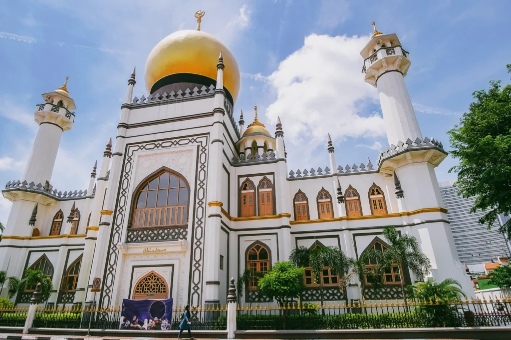 A low-angle, wide shot of the grand white facade of the Sultan Mosque (Masjid Sultan), highlighting its large golden dome, ornate minarets, and arched windows against a bright blue sky with light clouds.