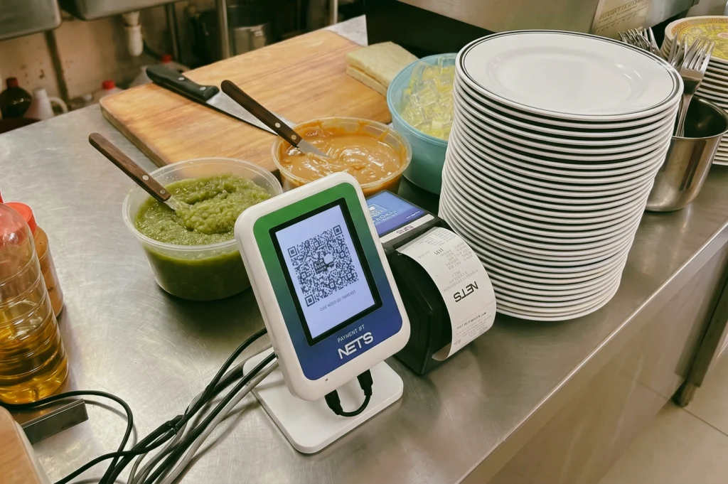 Traditional Singapore kopitiam prep station with kaya spread, butter and stacked plates beside NETS QR payment terminal – eye-level medium shot highlighting modern cashless coffee shop culture.