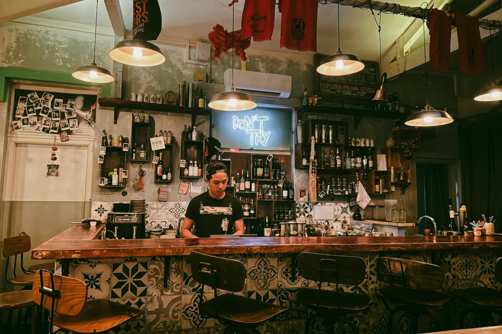 Eye-level front-facing shot of Sago House bartender preparing custom cocktails behind a rustic bar counter with shelves of liquor, neon sign, and eclectic décor in Duxton Hill Singapore