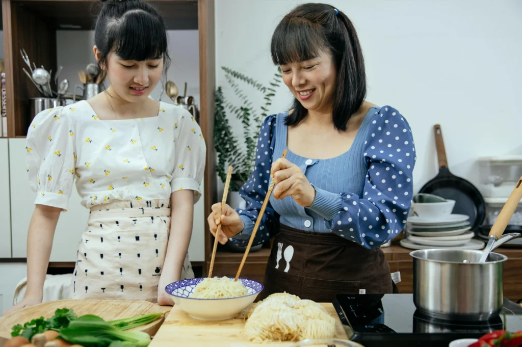 Eye-level medium shot of two people preparing fresh noodles together in a home kitchen, demonstrating traditional Singapore cooking techniques and the passing of generational recipes and food heritage
