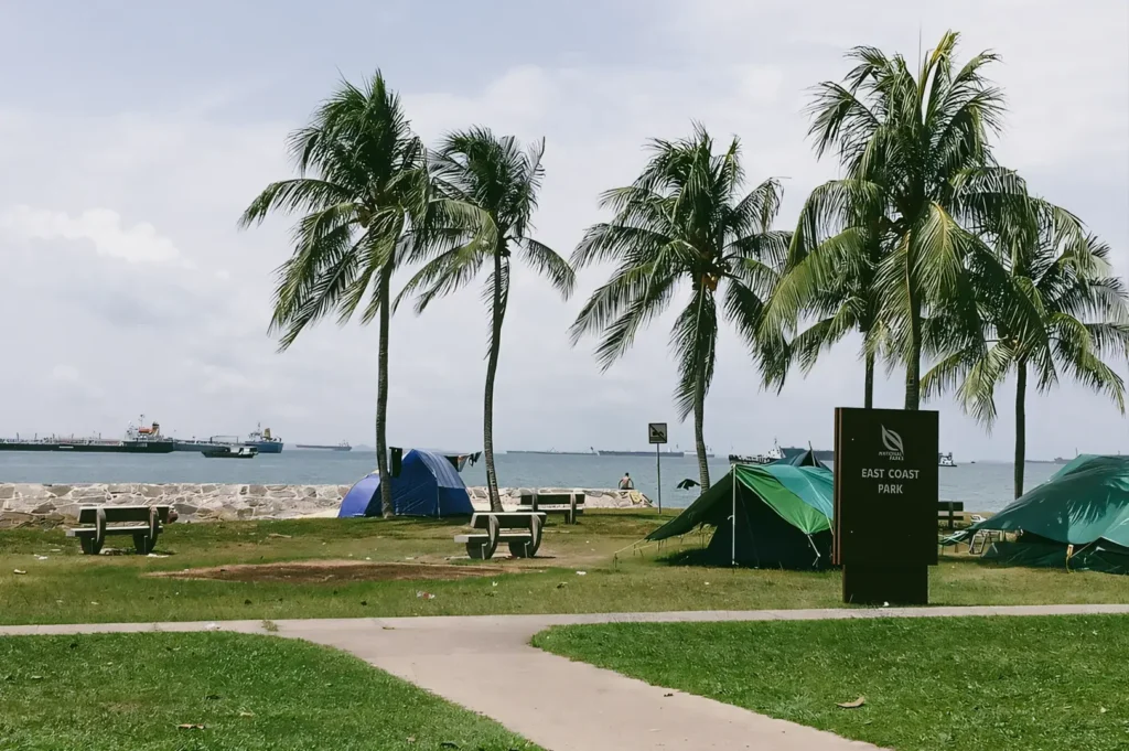 Eye-level wide shot of East Coast Park Singapore featuring palm-lined coastal park, camping tents, seaside area, and recreational spaces illustrating outdoor leisure and family-friendly public spaces.