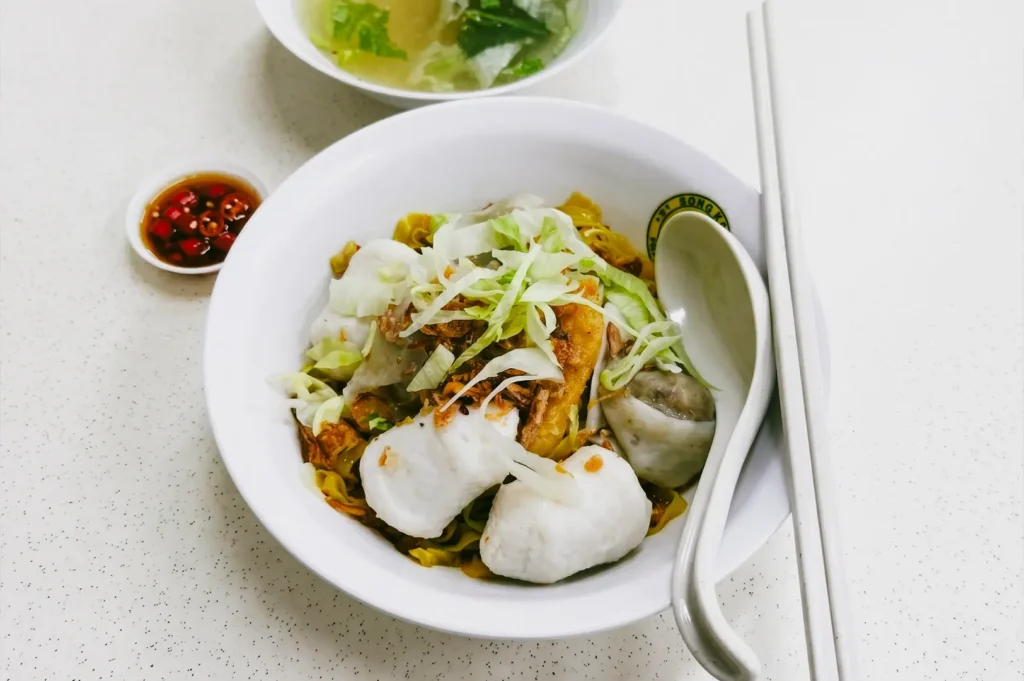 A top-down, close-up shot of a white ceramic bowl filled with handmade fishball noodles, shredded cabbage, and fried shallots on a speckled table, accompanied by a small side dish of red sliced chilis in soy sauce and white chopsticks.