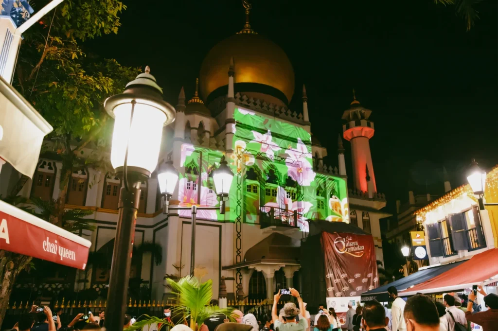 A low-angle night shot of the Sultan Mosque featuring a vibrant green and pink floral light projection mapping display across its white exterior walls, with crowds of people watching and street lamps illuminating the foreground.