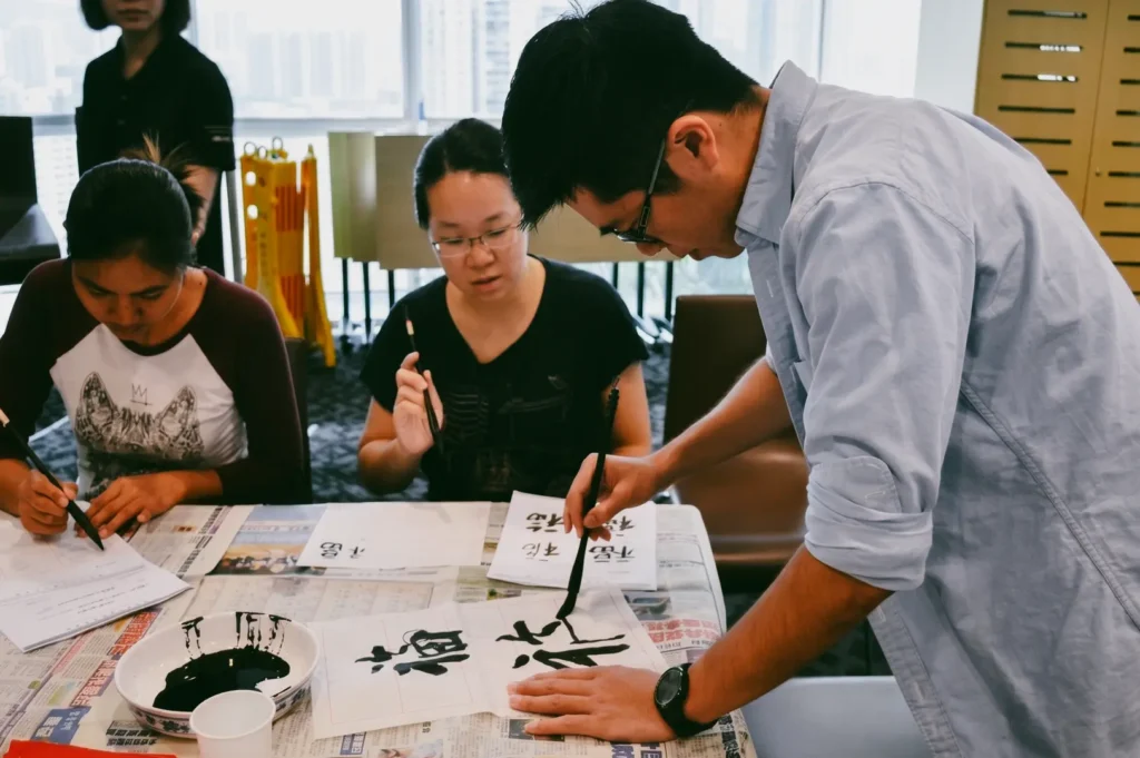 Over-the-shoulder medium shot angle of a calligraphy workshop with participants practicing Chinese brush writing and ink techniques, showcasing community learning, patience, and mindfulness.