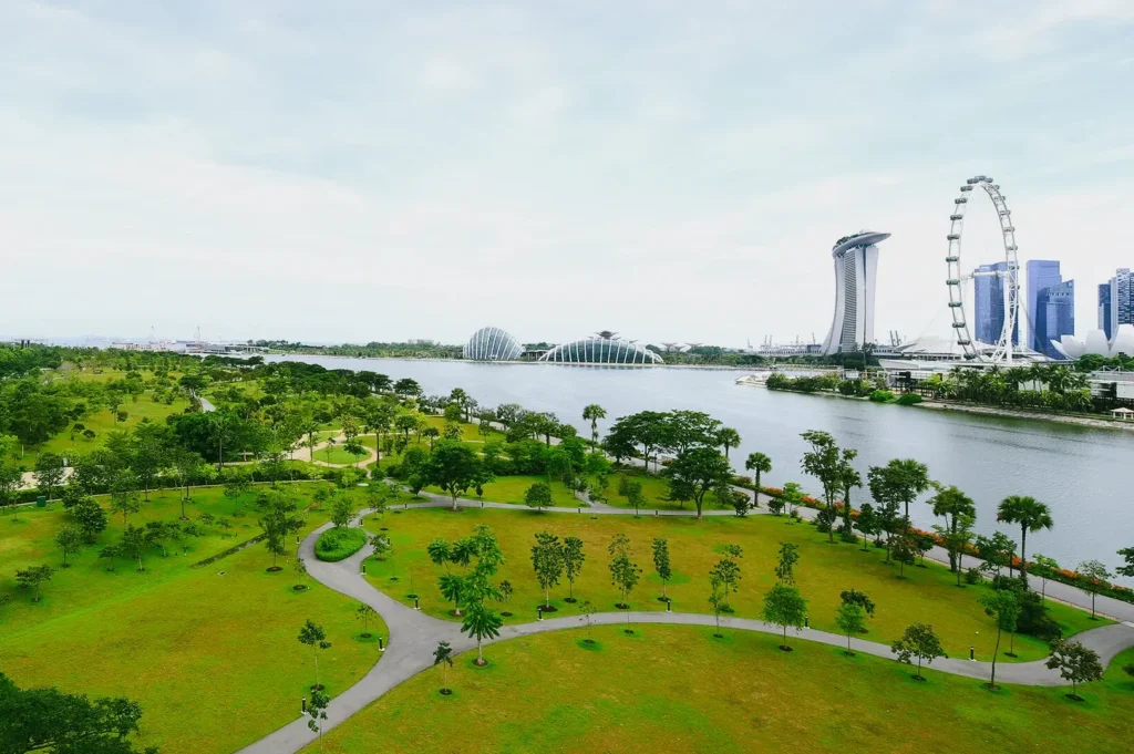 High-angle aerial panoramic shot of Bay East Garden Singapore with Marina Bay waterfront, Gardens by the Bay landscape, skyline views, and connected park paths showcasing expansive green spaces and urban planning.