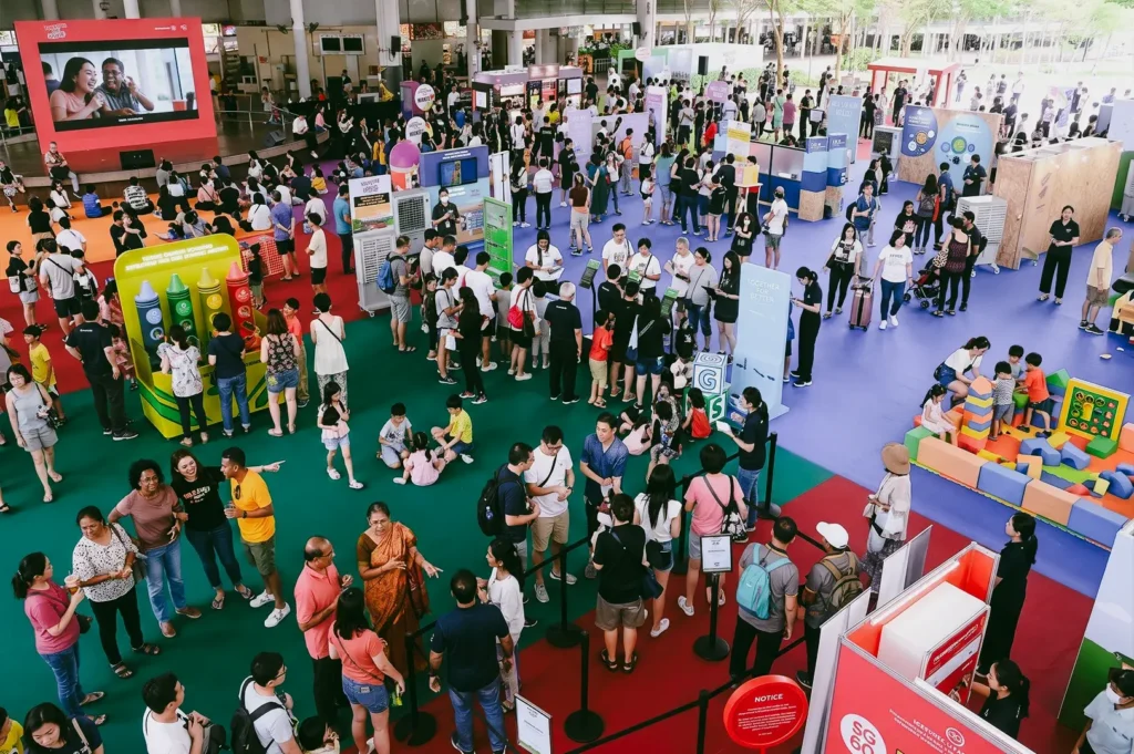 High-angle wide shot of a bustling community event in Singapore featuring crowded exhibition booths, families and residents interacting in colorful public space, highlighting real-world community engagement and social connection