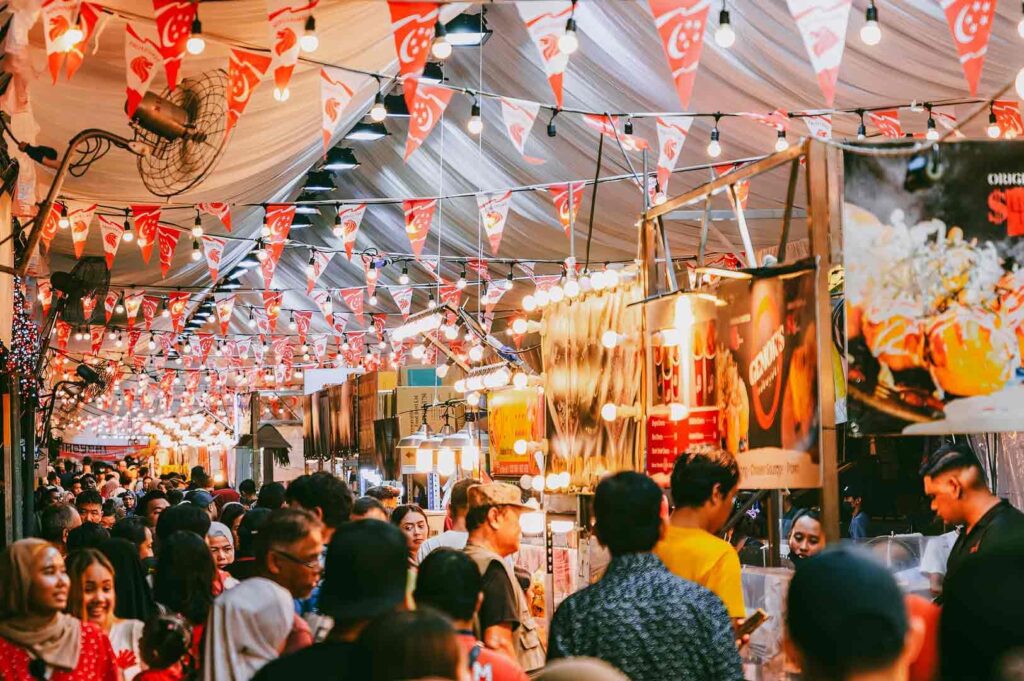 Eye-level wide-angle shot of a crowded Singapore pasar malam with rows of food stalls, string lights, and national flags above shoppers, showcasing vibrant community events, street food culture, and real-world social interaction