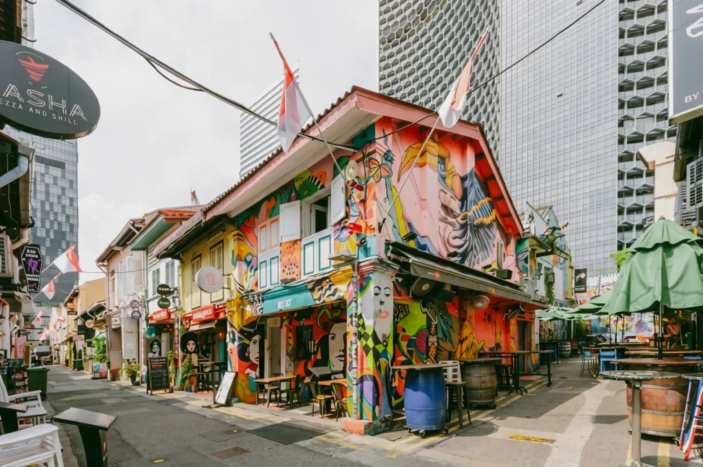 An eye-level, wide shot looking down Haji Lane, showcasing eclectic shophouses painted with bright, chaotic graffiti murals, outdoor cafe seating with green umbrellas, and modern skyscrapers rising in the background.