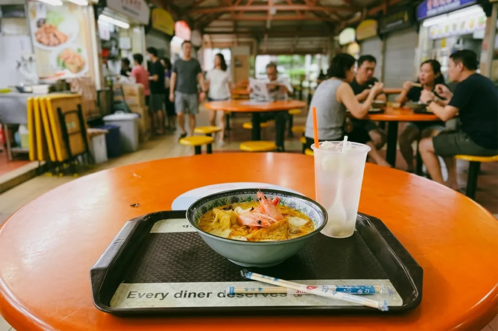 A low-angle, medium shot focusing on an orange table inside a hawker centre, holding a tray with a fresh bowl of laksa topped with prawns and a plastic cup of iced drink, while other diners sit blurred in the background.