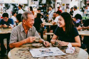 Eye-level medium shot of two people sharing coffee and conversation at a Singapore hawker centre, illustrating face-to-face interaction and meaningful real-world community connections in everyday life
