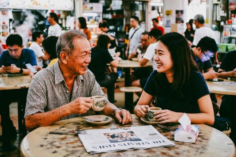 Eye-level medium shot of two people sharing coffee and conversation at a Singapore hawker centre, illustrating face-to-face interaction and meaningful real-world community connections in everyday life