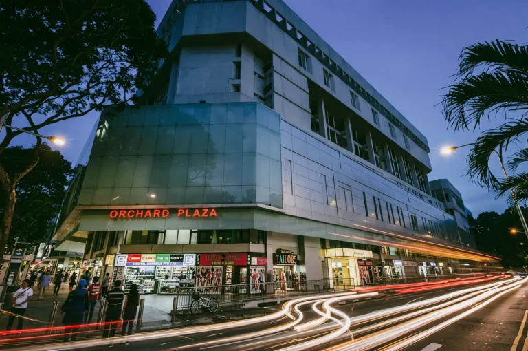 Wide-angle eye-level exterior view of Orchard Plaza building at dusk, featuring the illuminated ‘ORCHARD PLAZA’ sign above street-level shops, glass and concrete facade, pedestrians on the sidewalk, and long-exposure light trails from passing traffic creating bright streaks along the road under a dim blue evening sky.