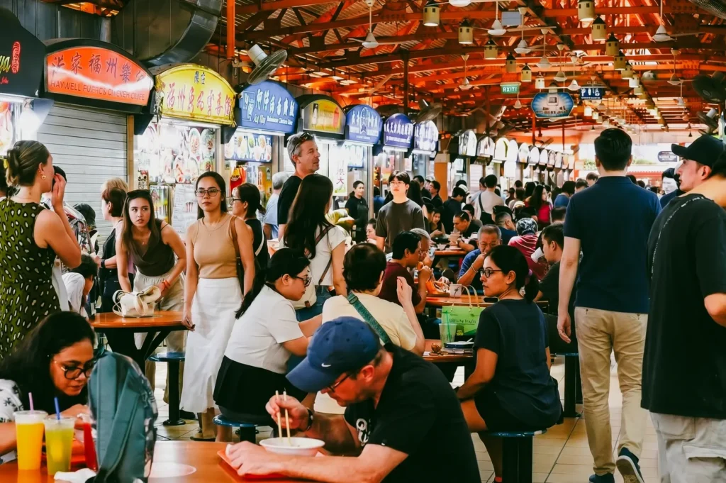 An eye-level, wide-angle shot highlighting a crowded communal dining area inside a brightly lit Singapore hawker centre, showing various local patrons sitting at tables and walking along the aisle next to food stalls under a high roof structure.