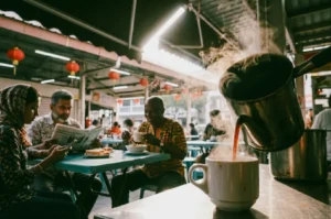 A three-quarter, close-up shot focusing on steaming dark coffee pouring from a metallic kettle into a white ceramic cup on a counter, with a blurry background showing a diverse group of locals conversing and reading newspapers at blue tables under red lanterns.