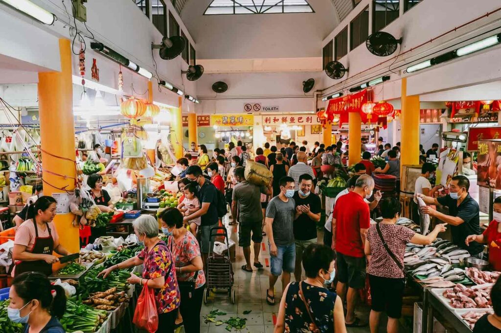Eye-level wide shot of a vibrant Singapore wet market with shoppers and vendors exchanging goods, showcasing local market culture, neighborhood life, and authentic community interaction