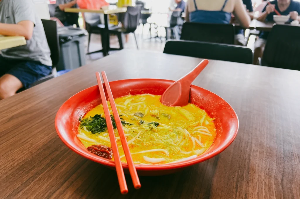 Close-up eye-level shot of a traditional Singapore laksa served in a red bowl at a hawker centre, featuring rich coconut curry broth, rice vermicelli noodles, tofu puffs, sambal chili, and fresh herbs on a dining table.
