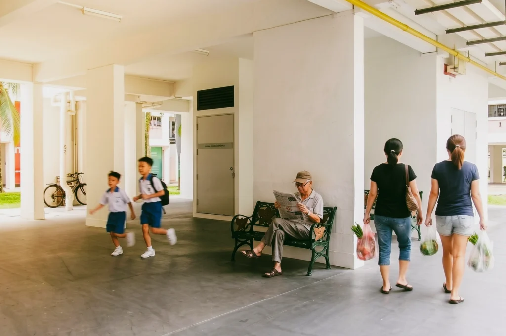 Wide-angle eye-level shot of a Singapore HDB void deck showing everyday community interaction, with residents walking, children passing through, and an elderly person reading on a bench, highlighting social architecture and shared neighbourhood spaces.