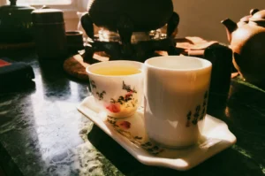 Low-angle close-up shot: Traditional Chinese tea cups filled with golden tea on a ceramic tray beside a teapot, illuminated by warm morning sunlight in a cozy tea ceremony setting, creating a serene dawn tea experience and mindful relaxation atmosphere