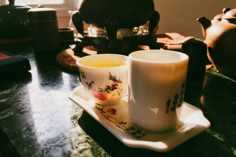 Low-angle close-up shot: Traditional Chinese tea cups filled with golden tea on a ceramic tray beside a teapot, illuminated by warm morning sunlight in a cozy tea ceremony setting, creating a serene dawn tea experience and mindful relaxation atmosphere