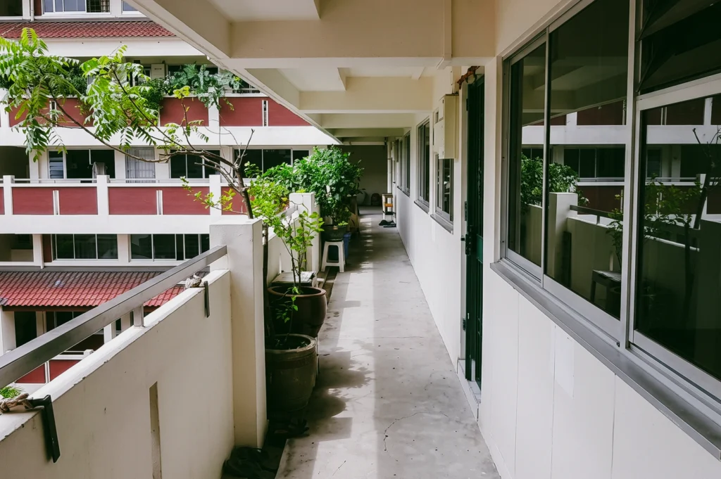 Leading lines perspective shot along a Singapore HDB corridor with potted plants and open walkways, emphasizing shared residential spaces, neighbour encounters, and everyday urban living design.