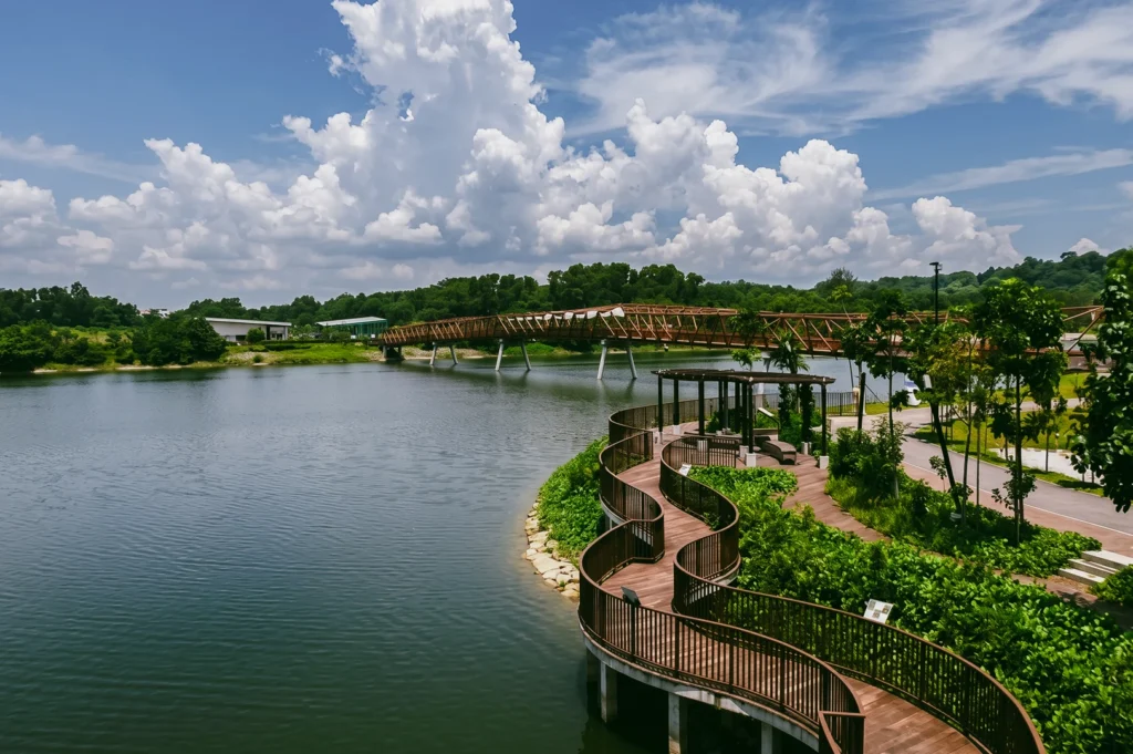 High eye-level wide-angle view of Punggol Waterway Park featuring a curved wooden boardwalk, landscaped greenery, and pedestrian bridge under dramatic skies, highlighting sustainable waterfront design and eco-friendly urban living in Singapore