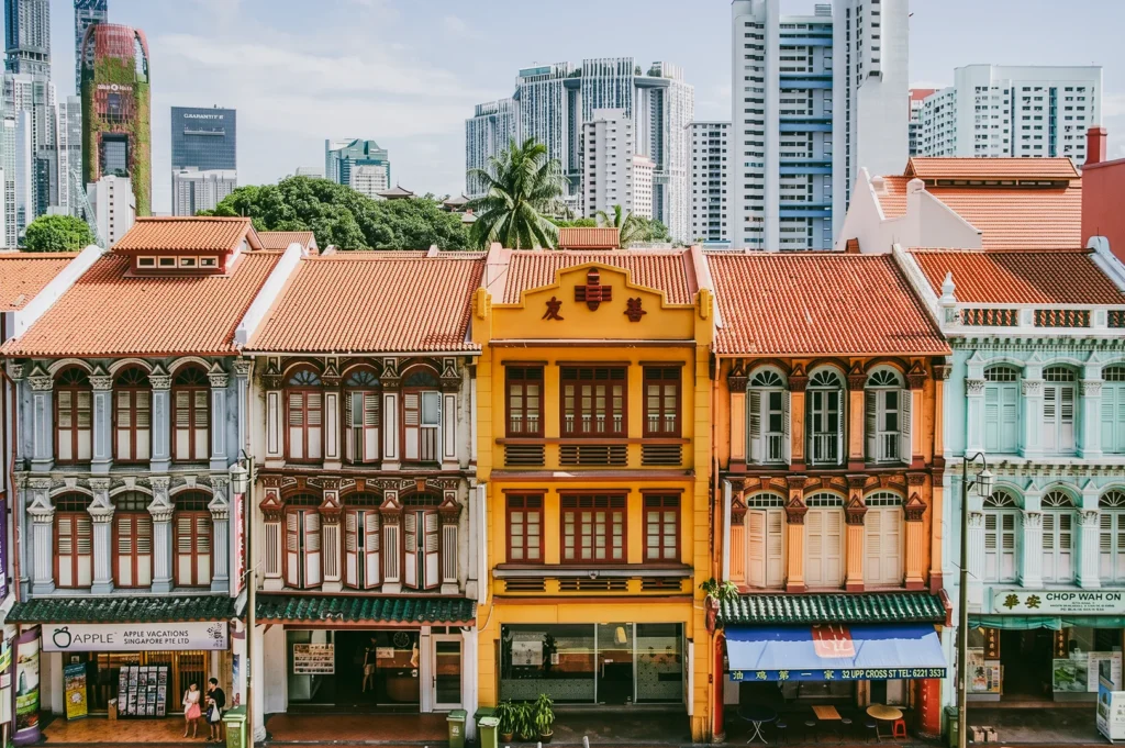 Eye-level wide-angle shot of colorful heritage shophouses in Singapore with modern high-rise buildings in the background, illustrating layered urban design and the coexistence of old and new neighbourhood architecture.
