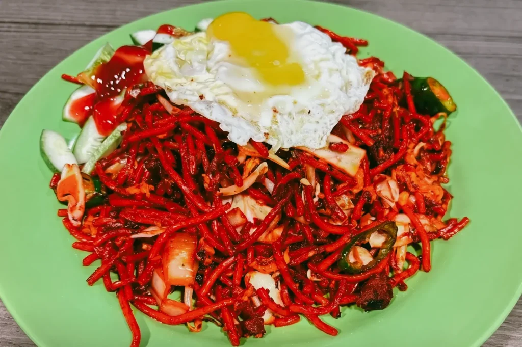 Top-down, high-angle shot of a full plate of vibrant red mee goreng noodles served on a bright green plastic hawker plate, topped with a fried egg featuring a slightly runny yolk, accompanied by sliced fresh cucumbers and a drizzle of sweet chili sauce on the side.