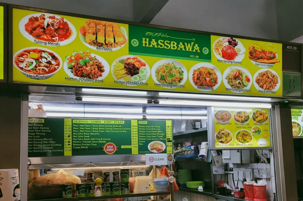Eye-level shot focusing closely on a brightly lit, yellow and green overhead menu board for the iconic Hass Bawa hawker stall, showcasing vivid photo listings and prices for traditional dishes like mee kuah, roti john, and traditional mee goreng variants above the food preparation area.