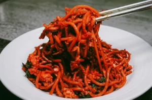 Close-up, eye-level shot of a heap of bright, reddish-orange fried mee goreng Singapore noodles piled high on a white rimmed plate, with metal chopsticks lifting a large bundle of noodles directly toward the camera, highlighting the glossy sauce, flecks of egg, and green vegetables