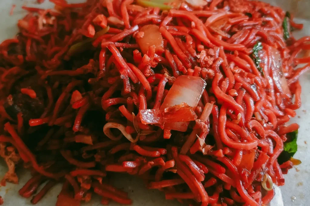 Macro, high-angle shot focusing tightly on a dense serving of red hawker-style fried noodles mixed with translucent pieces of cooked onion, cabbage, and green leafy vegetables, showcasing the moist, rich texture of the seasoned dish up close.